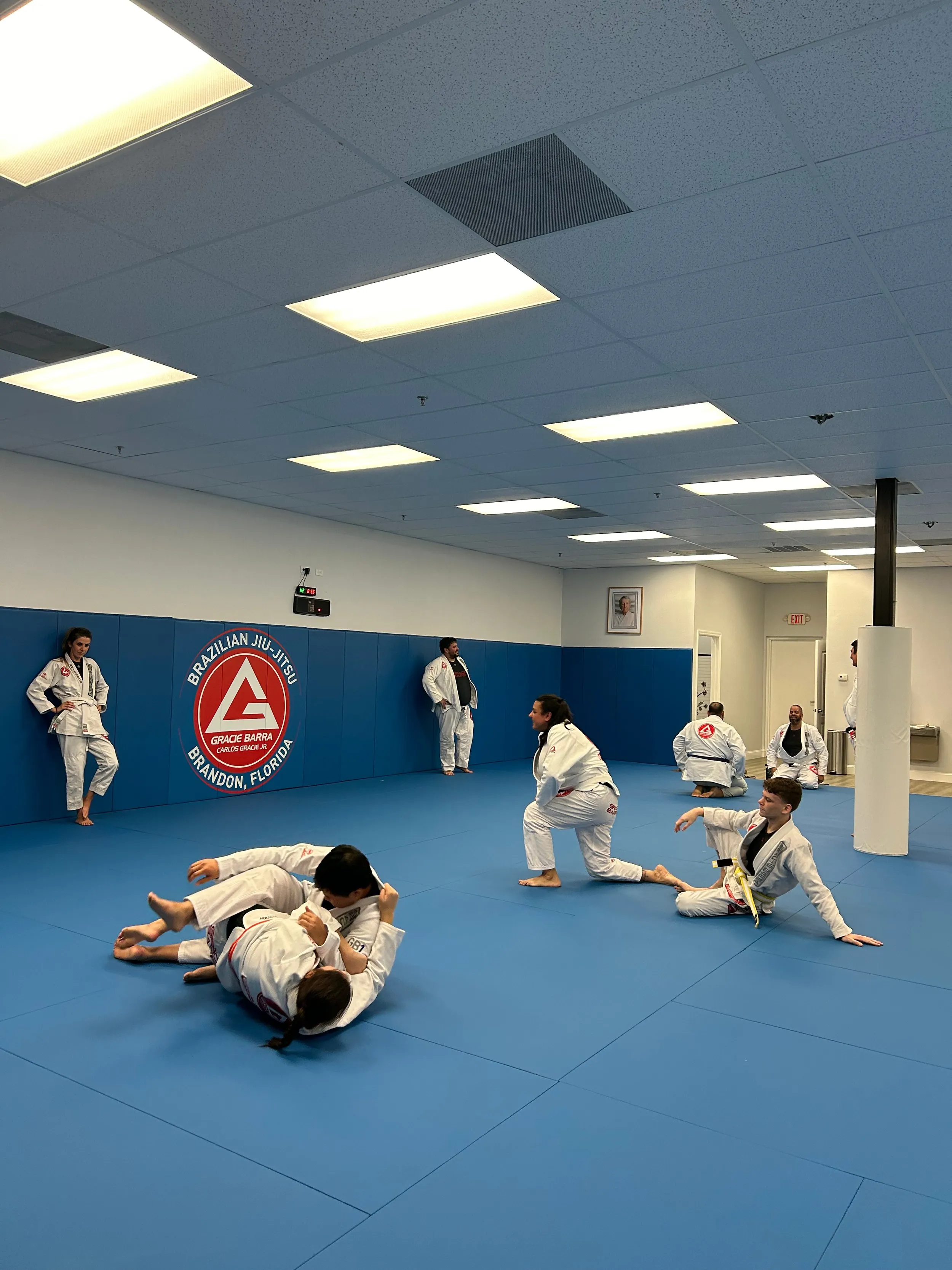 A Gracie Barra Brandon class in progress: students drilling in pairs on the blue mats while others rest and watch, under the academy’s wall crest.